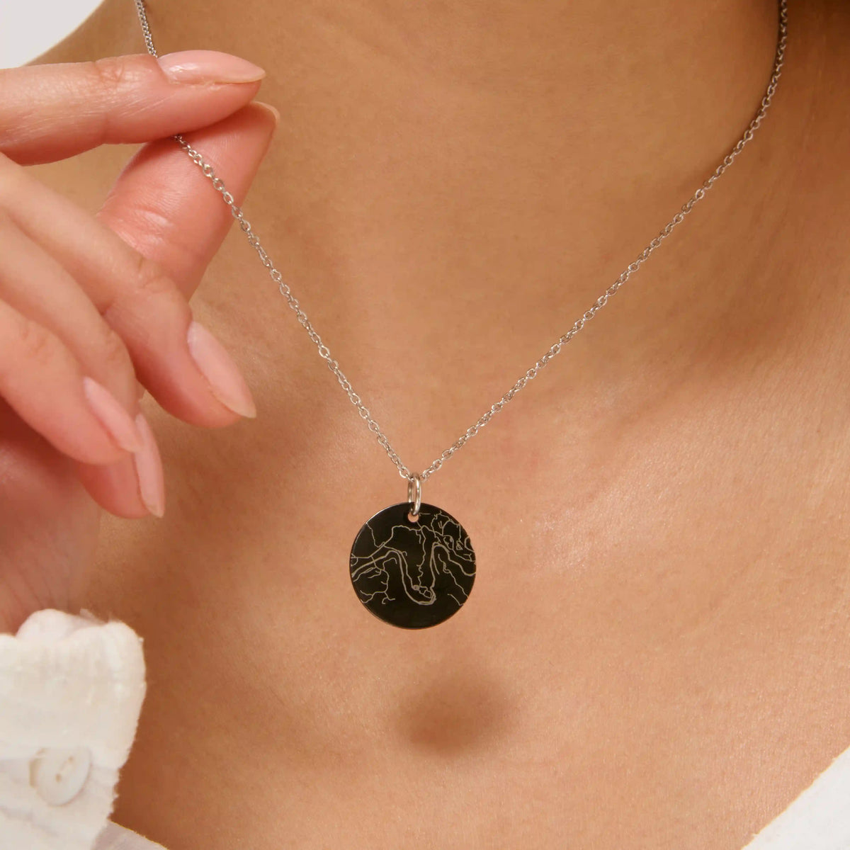 Black street map necklace engraved in silver lines, held between fingers at the neck