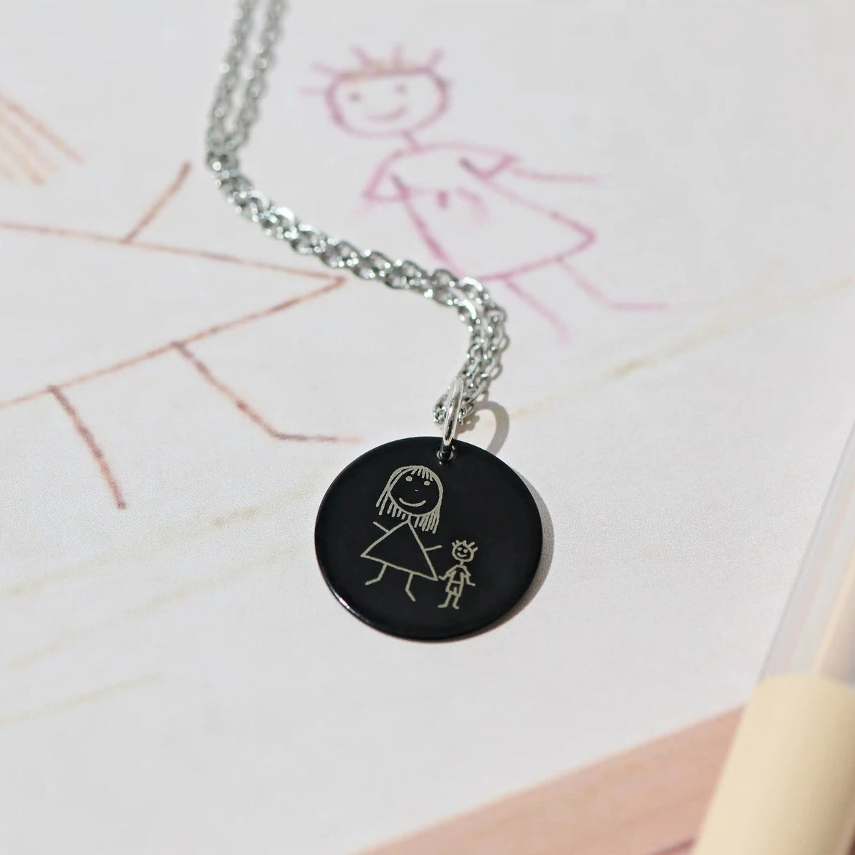 Black pendant engraved with a child's drawing, resting on the original picture