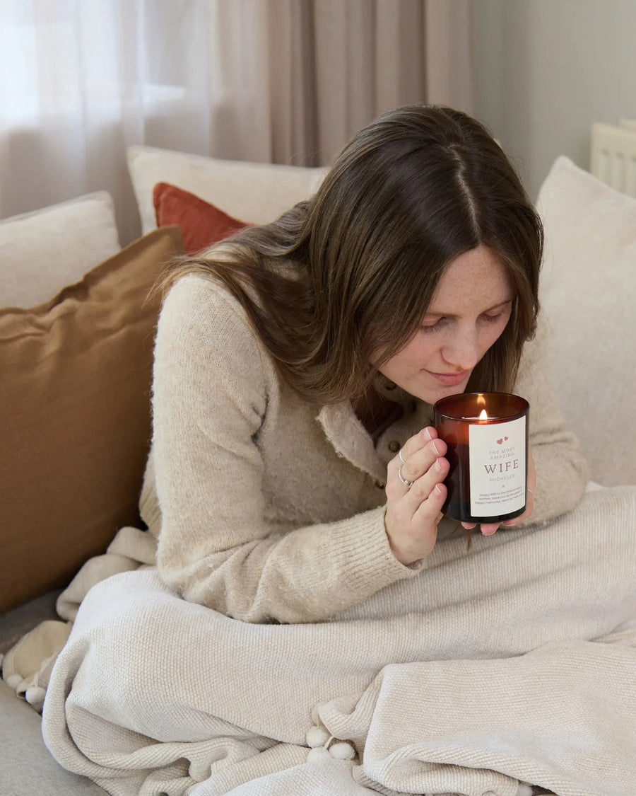 Woman wrapped in a blanket on a sofa, breathing in the scent of a personalised candle