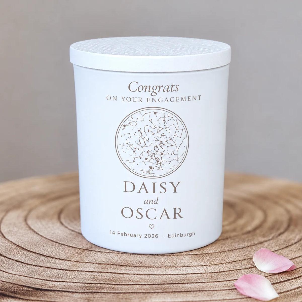 Engraved star map candle in a white glass jar, engagement design personalised with Daisy and Oscar with a congratulations message, on a wooden slice with petals